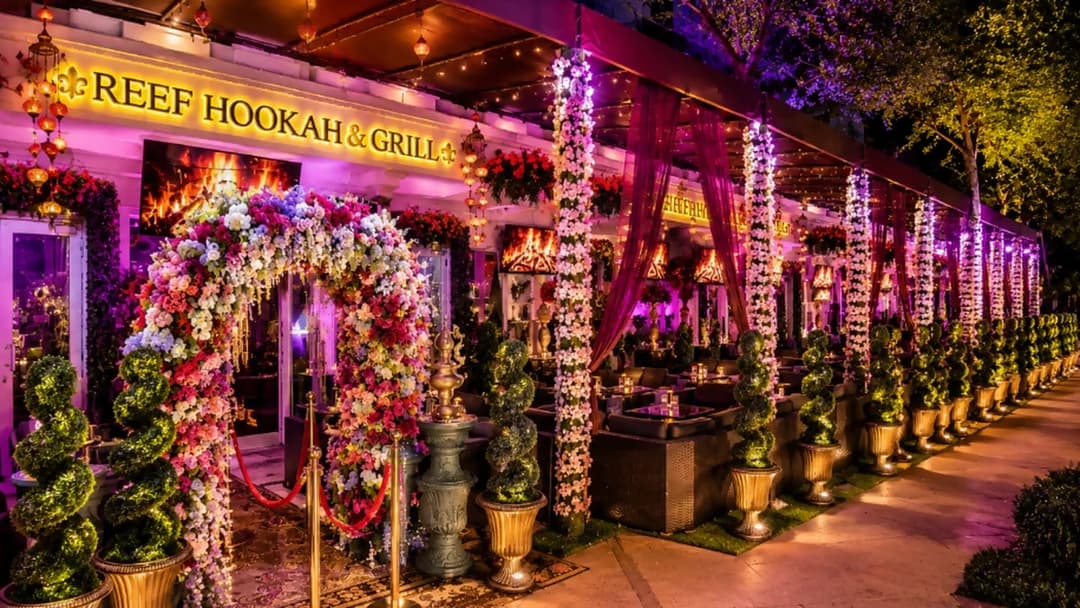 The entrance of Reef Hookah and Grill at night: hanging lanterns under a dark canopy ceiling, the gold REEF HOOKAH and GRILL sign, a fireplace screen above a floral archway with a red velvet rope